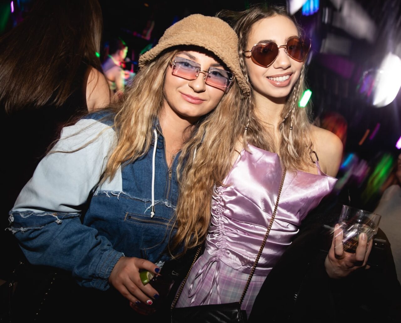 Two women in Y2K-inspired outfits and tinted sunglasses posing with drinks at a 2000’s Throwback Party.