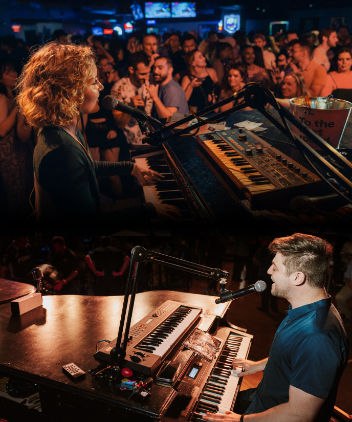Two live musicians perform dueling pianos on stage at a crowded bar, singing into microphones while playing keyboards as an energetic audience watches and dances.